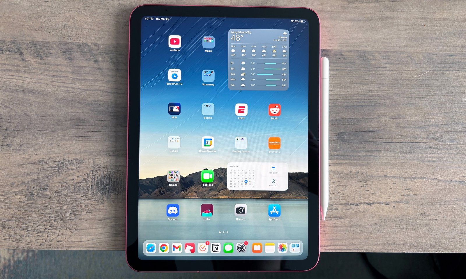 Apple iPad (A16) com lápis de maçã conectado ao lado, localizado em uma mesa de madeira marrom clara. Sua tela está ligada, uma exibição de uma tela inicial cheia de aplicativos e necessidades.