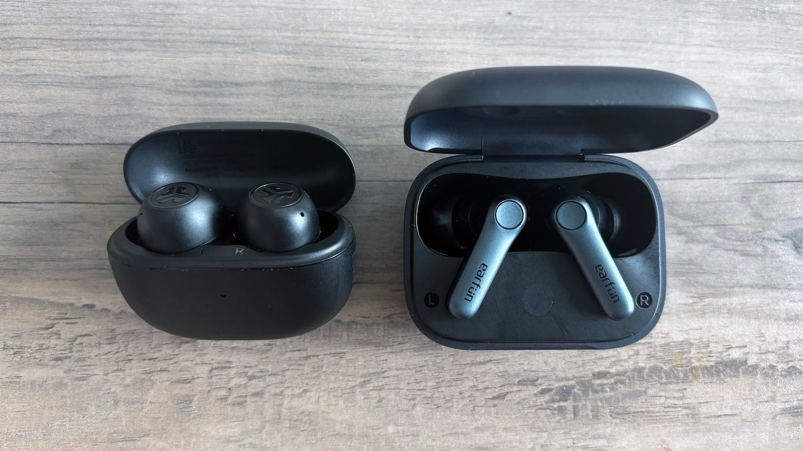 Dois casais negros de fones de ouvido sem fio, Jlab Go Pop ANC e Earfun Air Pro 4, descansam em uma mesa de madeira marrom clara com a abertura de suas caixas e expondo fones de ouvido durante o envio.